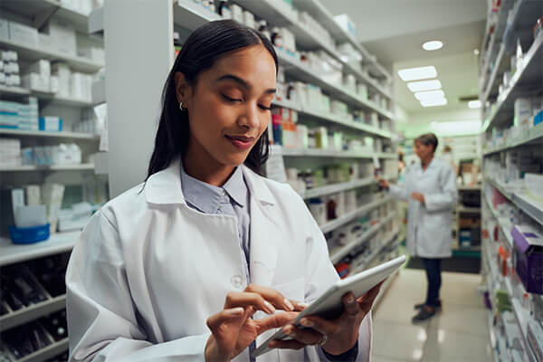 Smiling young female worker in pharmacy wearing labcoat checking inventory using digital tablet