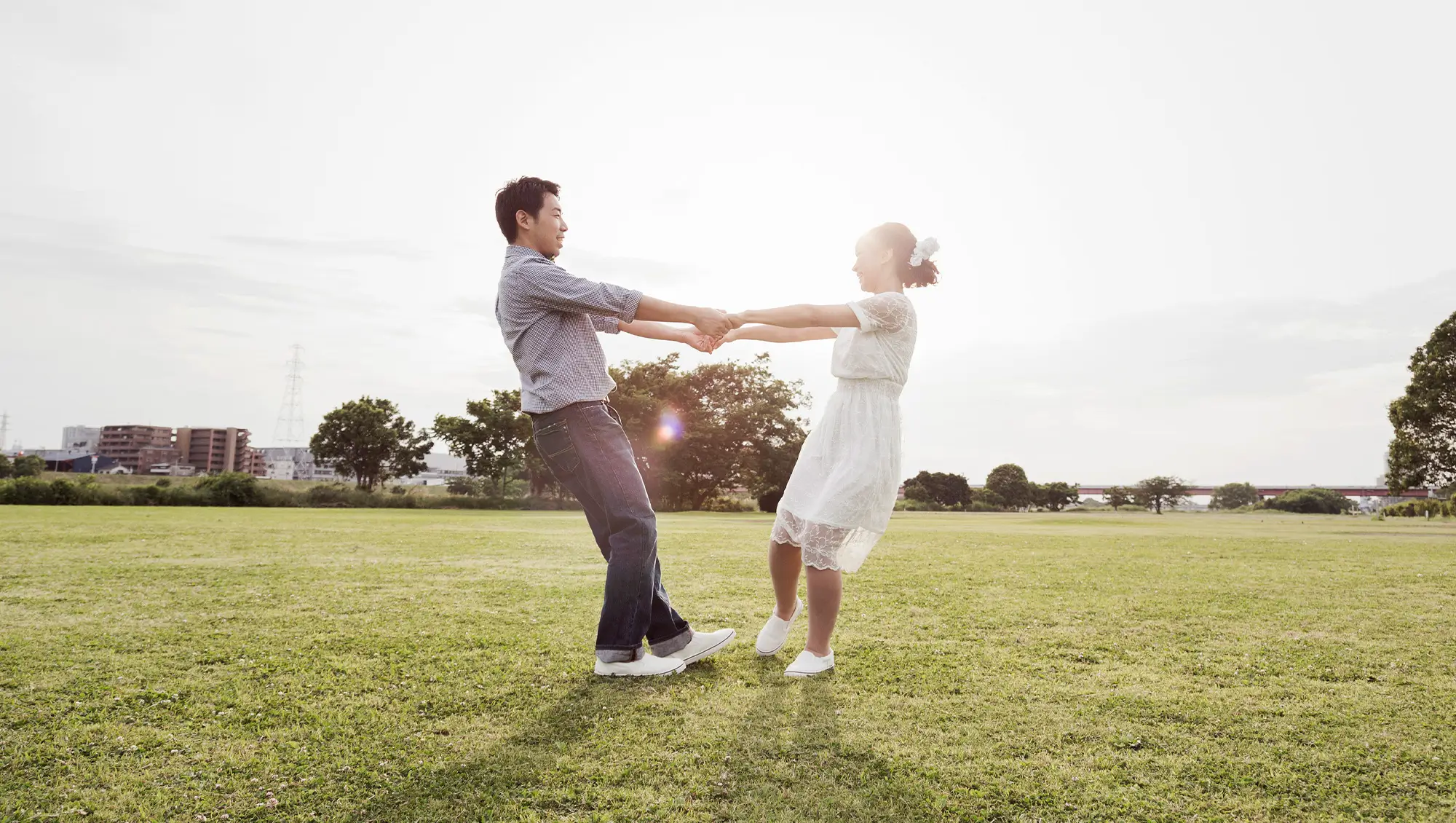 A couple holding hands and spinning together in a sunny park field, with the man wearing a striped shirt and jeans, and the woman in a white lace dress